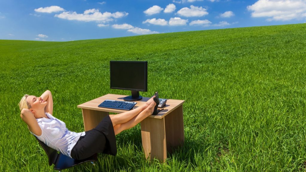 Mujer relajada con los pies sobre el escritorio y de fondo un paisaje con pasto verde y cielo azul
