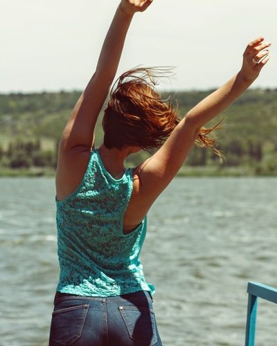 Mujer joven de espalda con manos al aire frente al lago